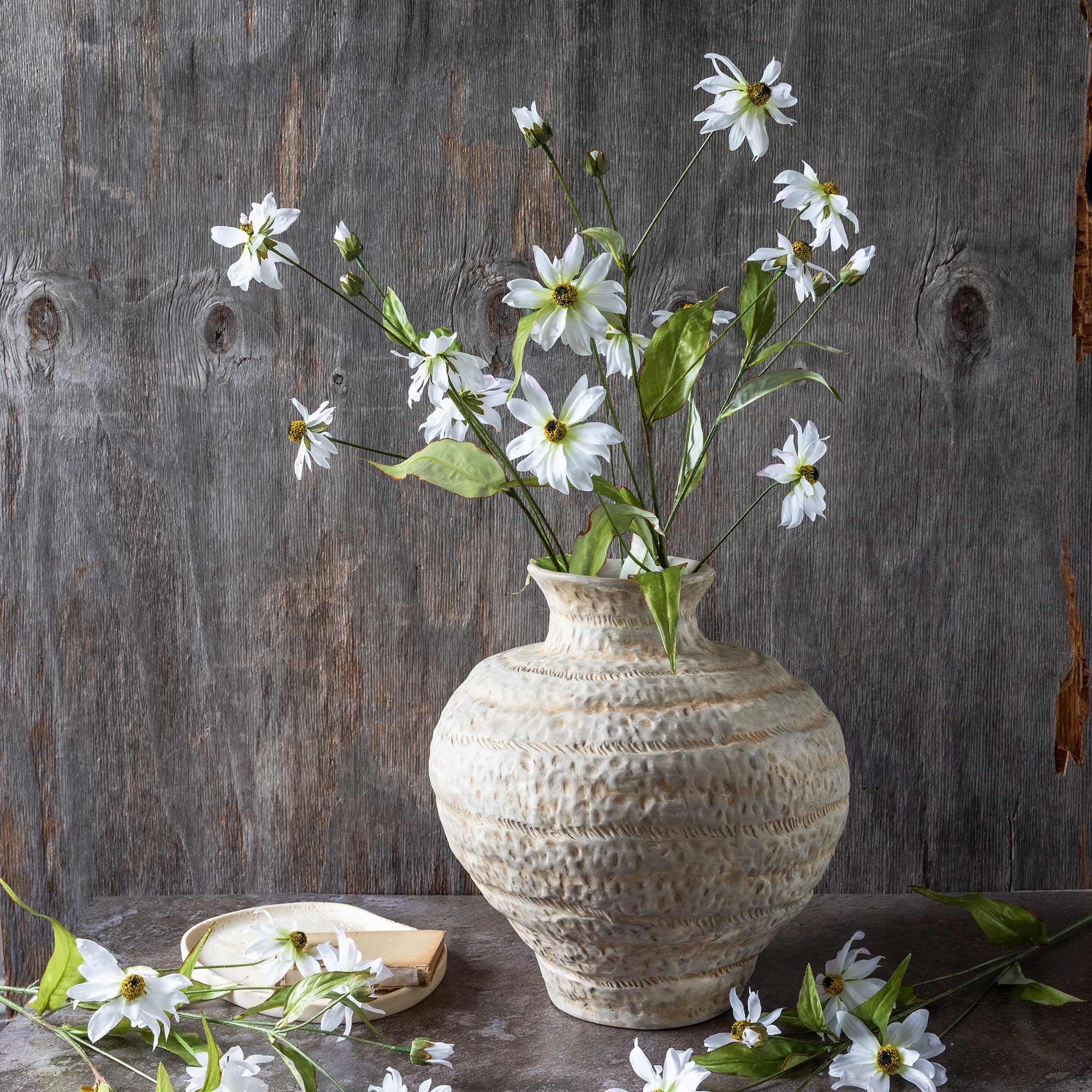 Faux cosmos arranged in a wide ceramic vase against a rustic wood backdrop