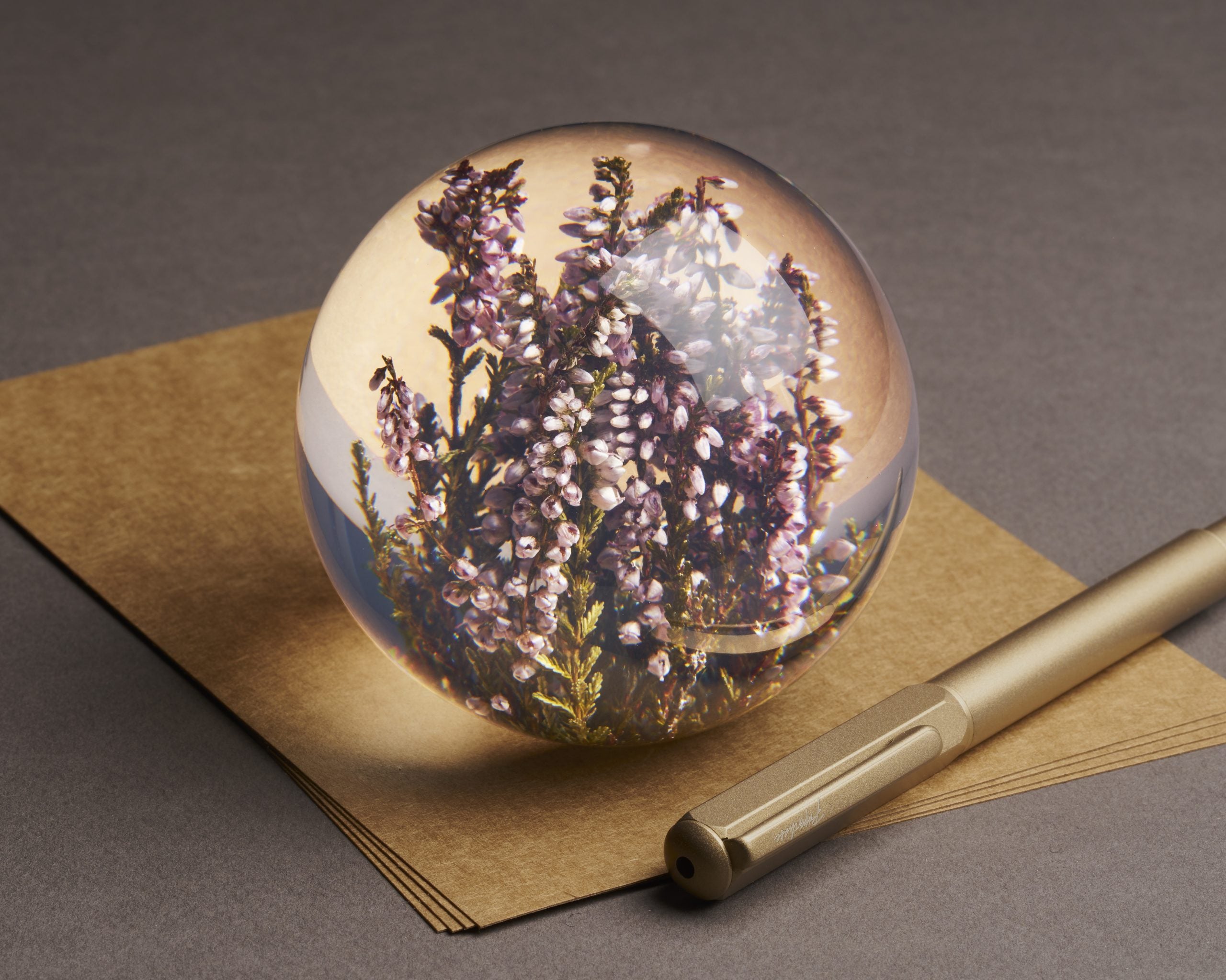 Botanical glass paperweight containing purple heather flowers resting on kraft paper beside a gold pen