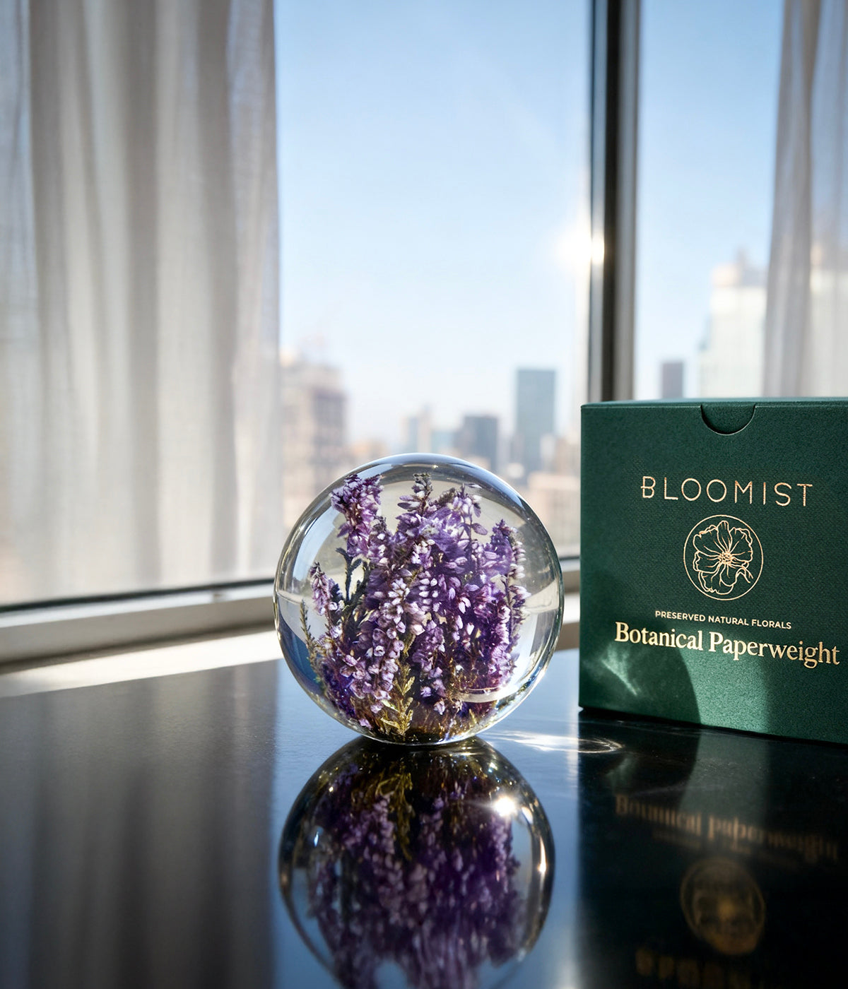 Botanical glass paperweight featuring purple heather flowers placed on a glossy surface beside a green Bloomist box, with a city view in the background