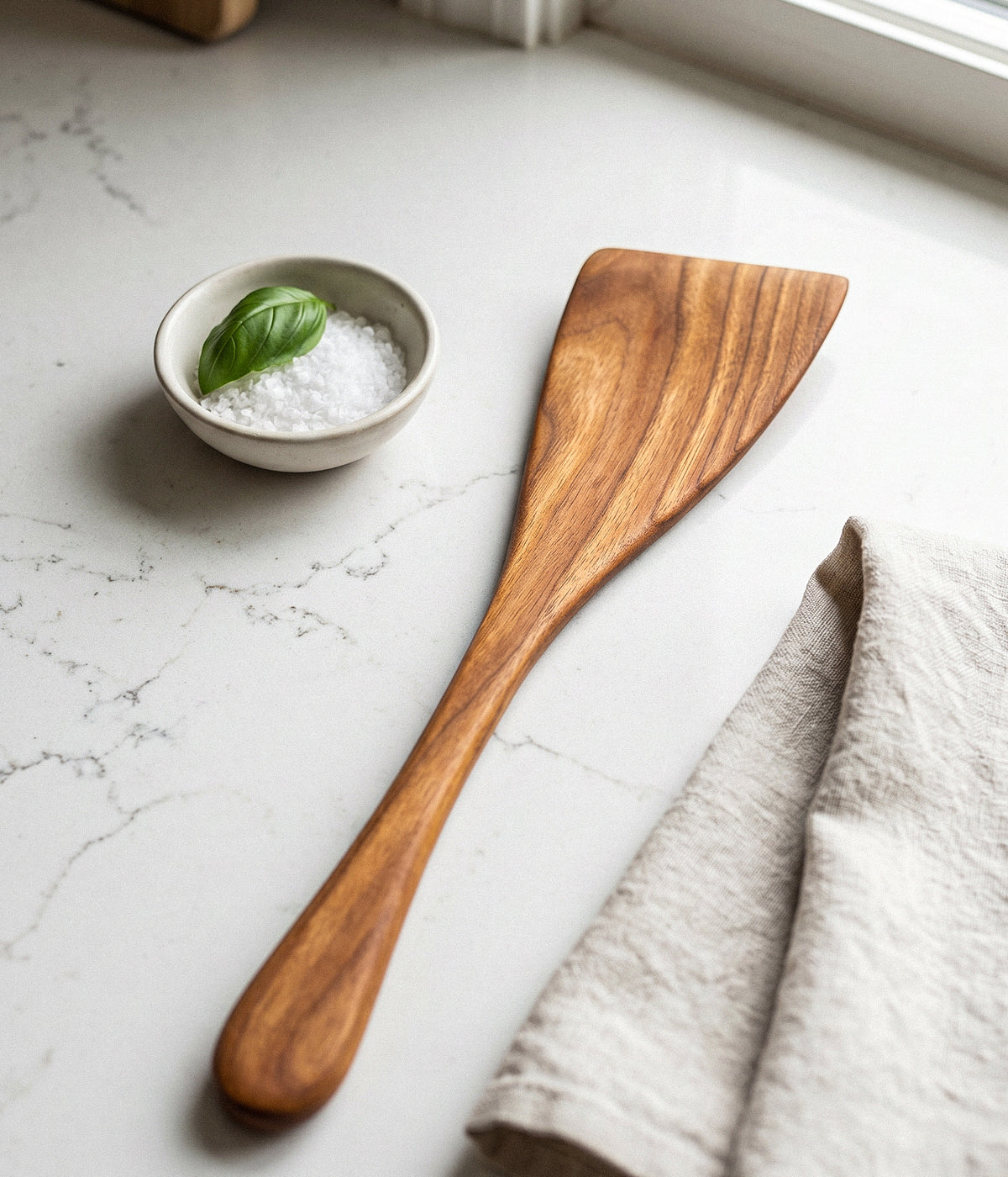 Wooden angled spatula on a marble surface with a small bowl of salt and basil leaves.