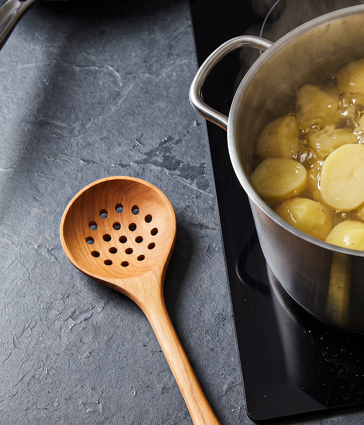 Wooden spoon with holes next to a pot of boiling potatoes on a dark surface