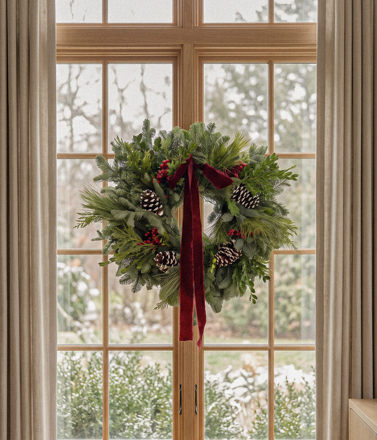 Margaux fresh fir wreath with burgundy velvet ribbon, red berries, and pine cones hanging on a wooden-framed glass door